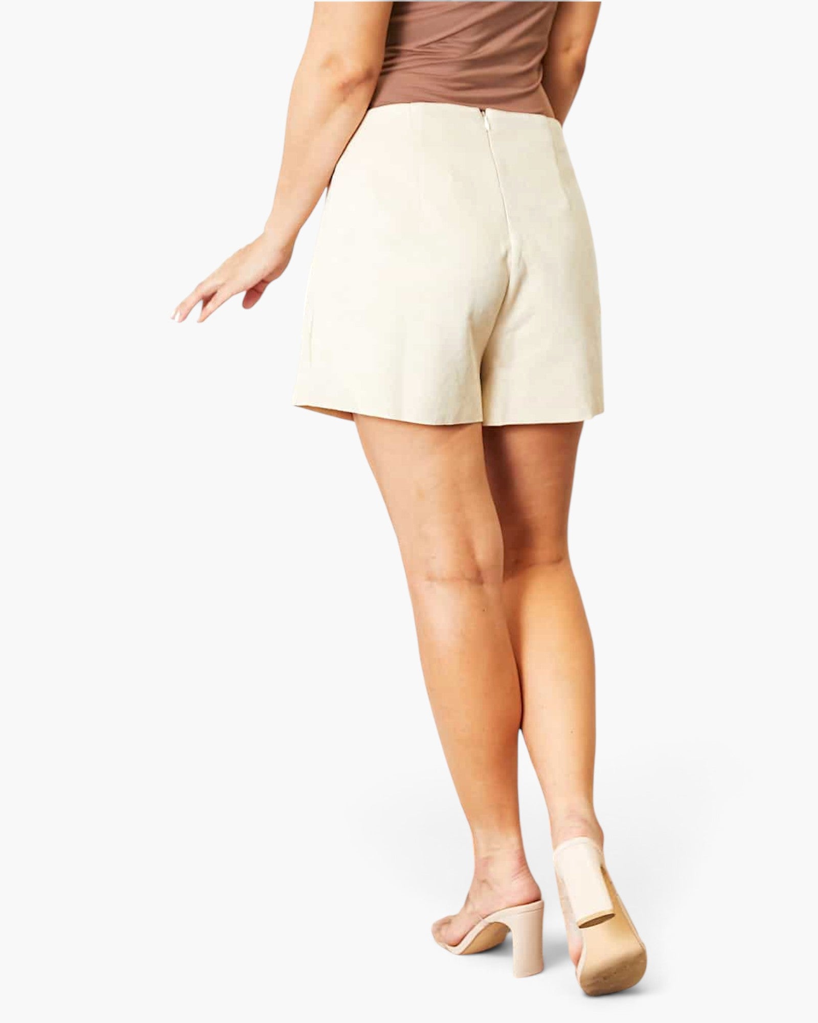 A model from the back wearing high-waisted, off-white shorts, a brown top, and beige high-heeled sandals against a plain white background.