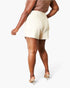 A model shown from the back wearing high-waisted cream shorts, a brown top, and clear-strap high heels against a light gray studio background.