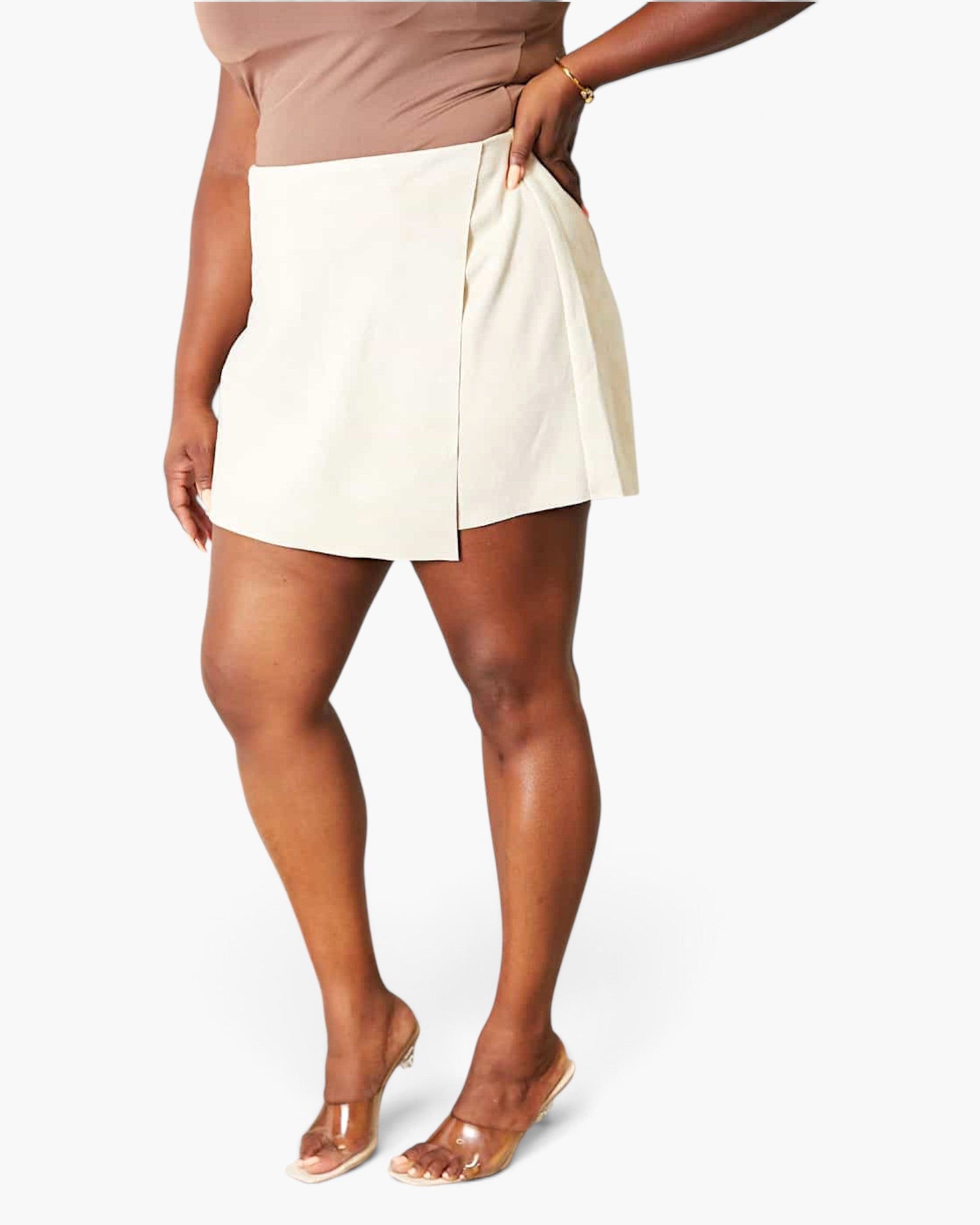 A model wearing a cream-colored wrap-style mini skirt with a pleated front detail, paired with a taupe top and clear high-heeled sandals, set against a plain light gray studio background.