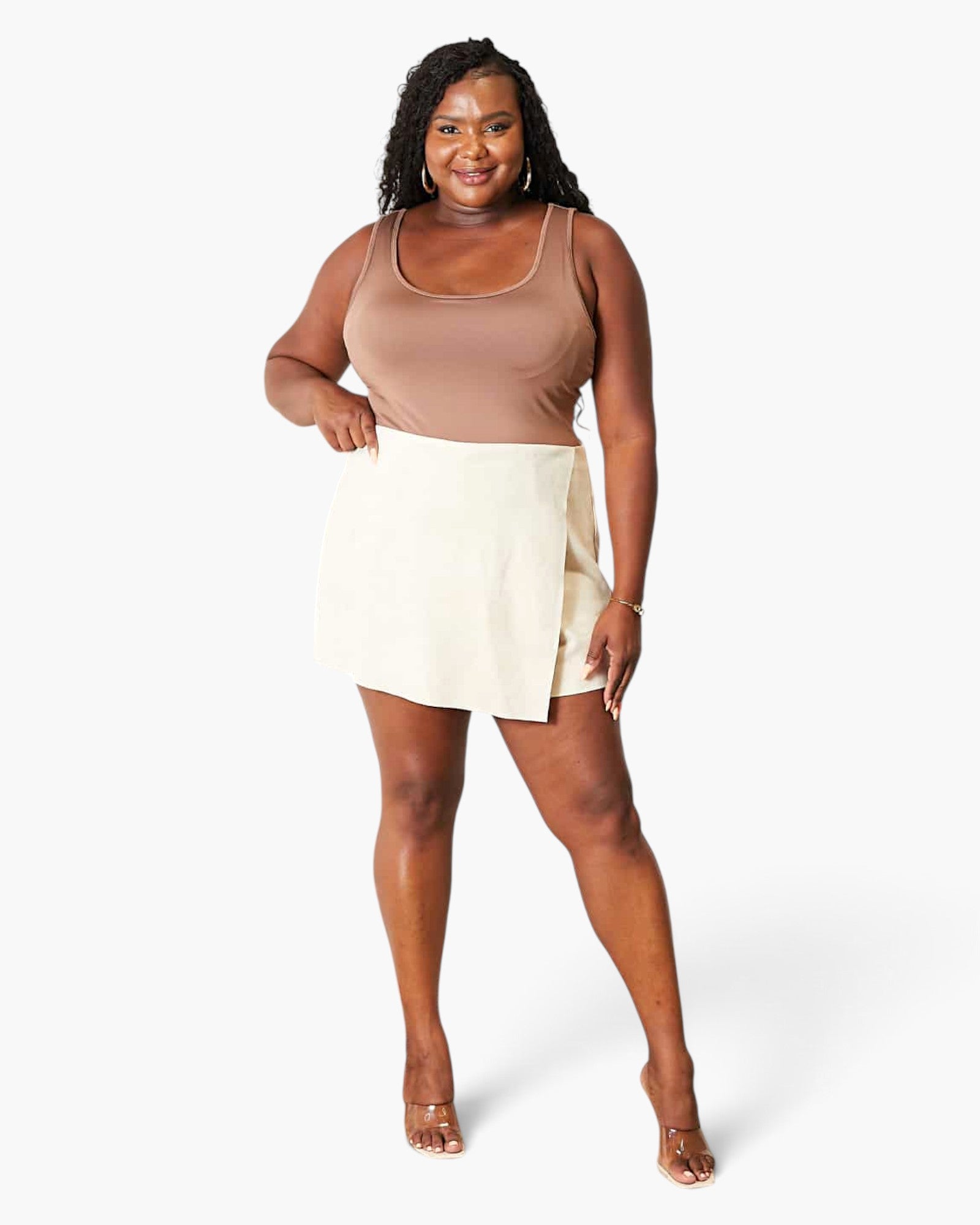 A woman modeling a sleeveless scoop-neck top in taupe and a cream mini skirt, paired with clear strap high-heeled sandals, against a solid white studio background.