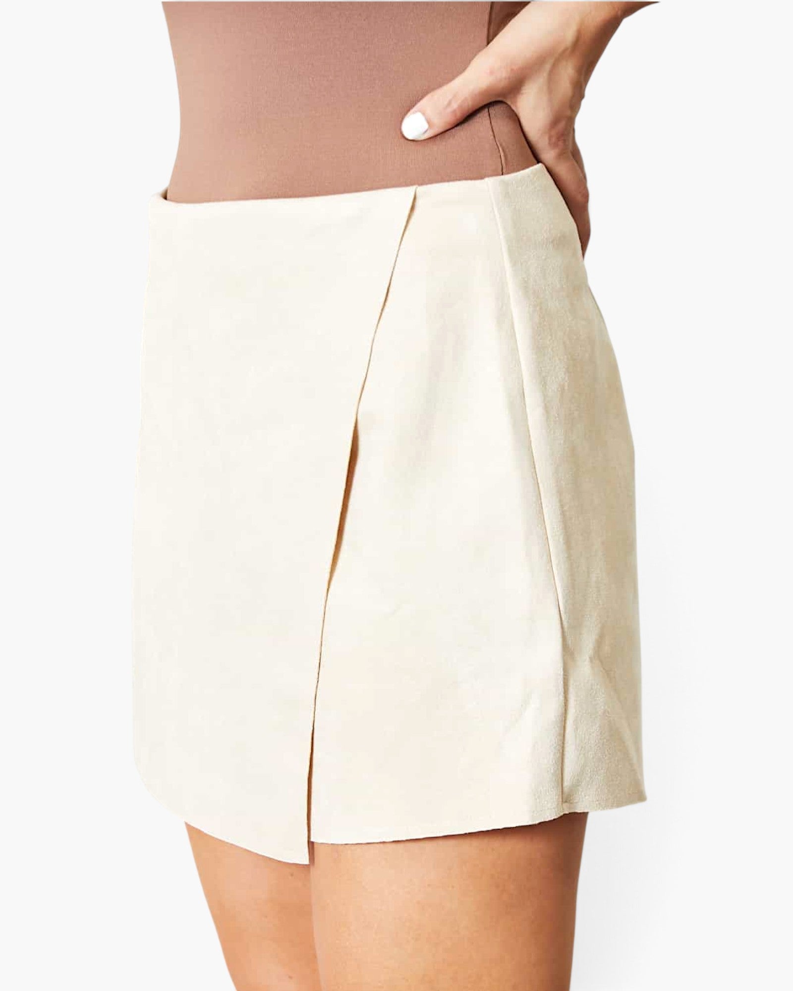 A close-up of a model wearing a light beige wrap-style mini skirt with a front slit, paired with a matching beige bodysuit against a plain white background.