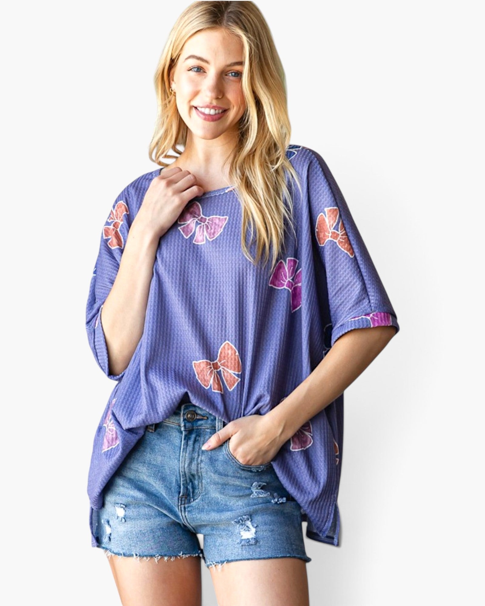 A woman modeling a purple waffle-knit short-sleeve top with a pink and orange bow print, paired with distressed denim shorts, against a plain white background.