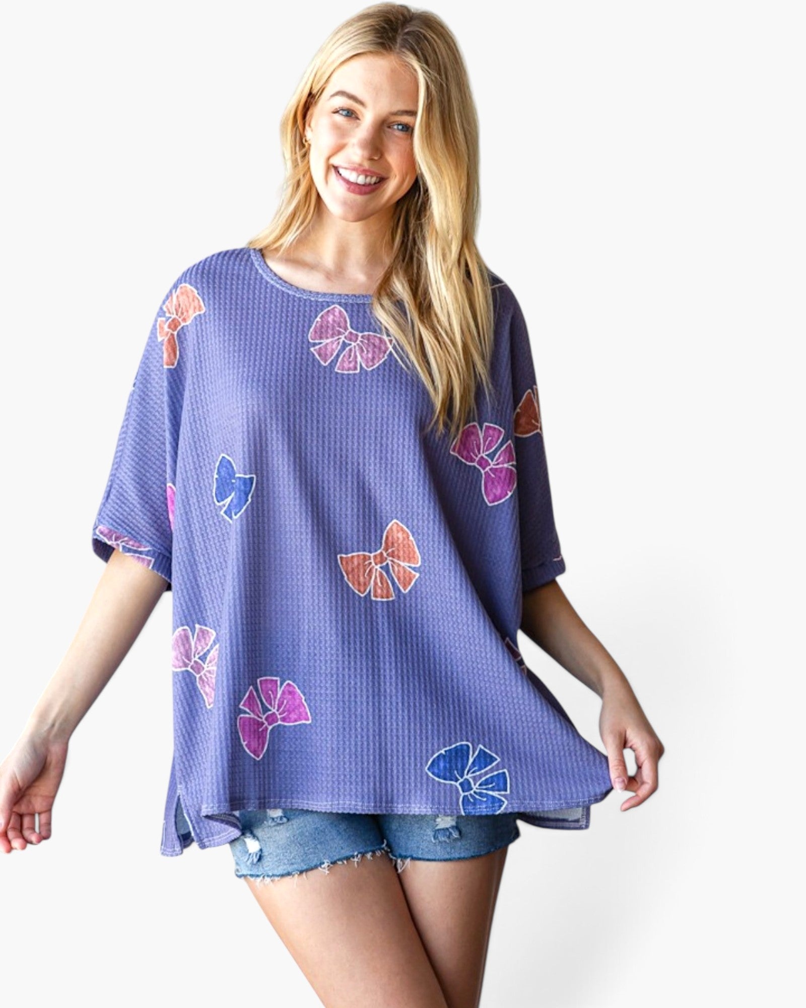 A woman modeling a short-sleeve waffle-knit top in lavender purple with colorful butterfly prints in pink, orange, and blue, paired with denim shorts against a plain white background.