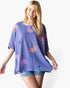 A woman modeling a short-sleeve waffle-knit top in lavender purple with colorful butterfly prints in pink, orange, and blue, paired with denim shorts against a plain white background.