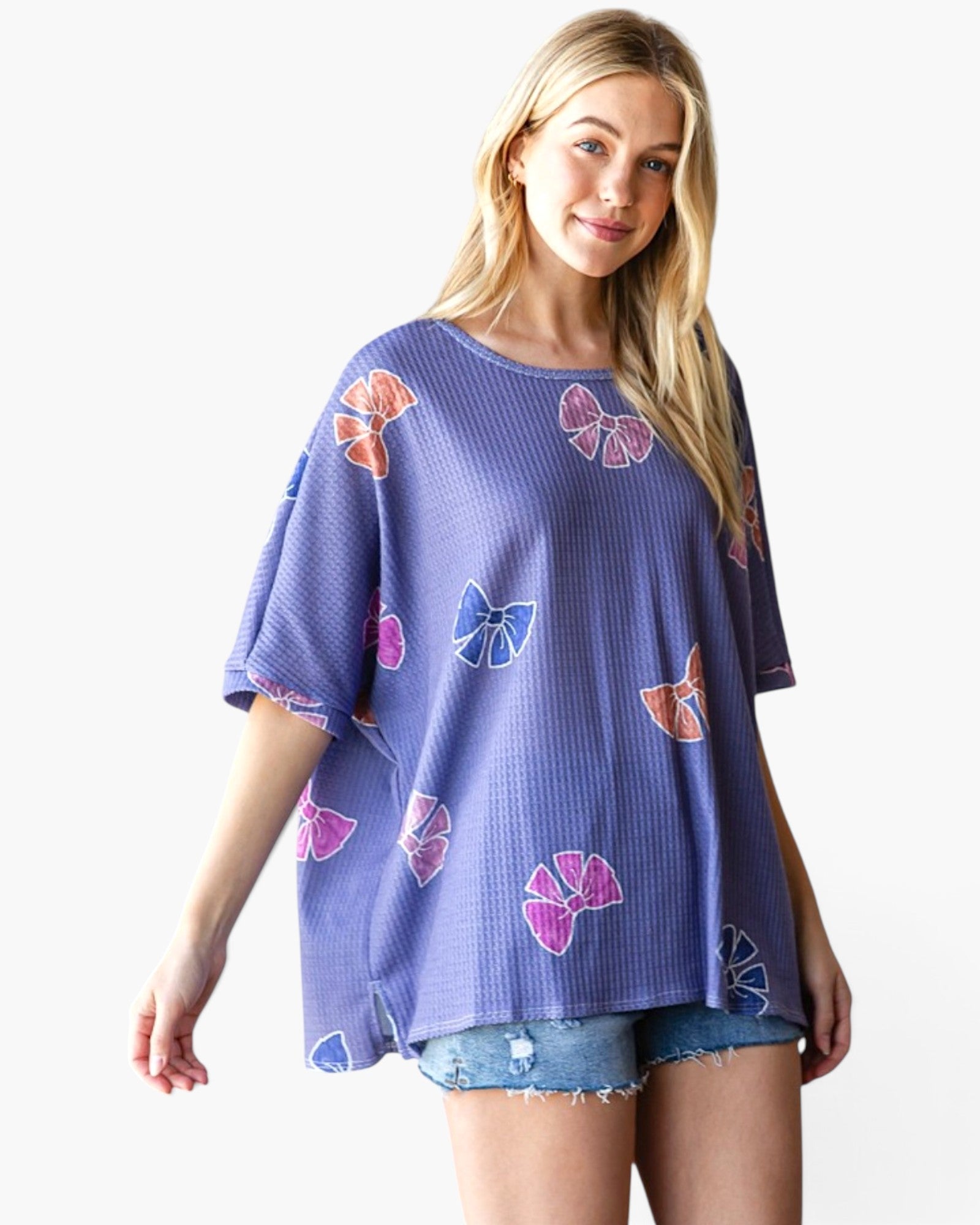 A woman modeling a lavender-purple waffle-knit short-sleeve top with colorful butterfly prints, paired with denim shorts, against a plain white background.