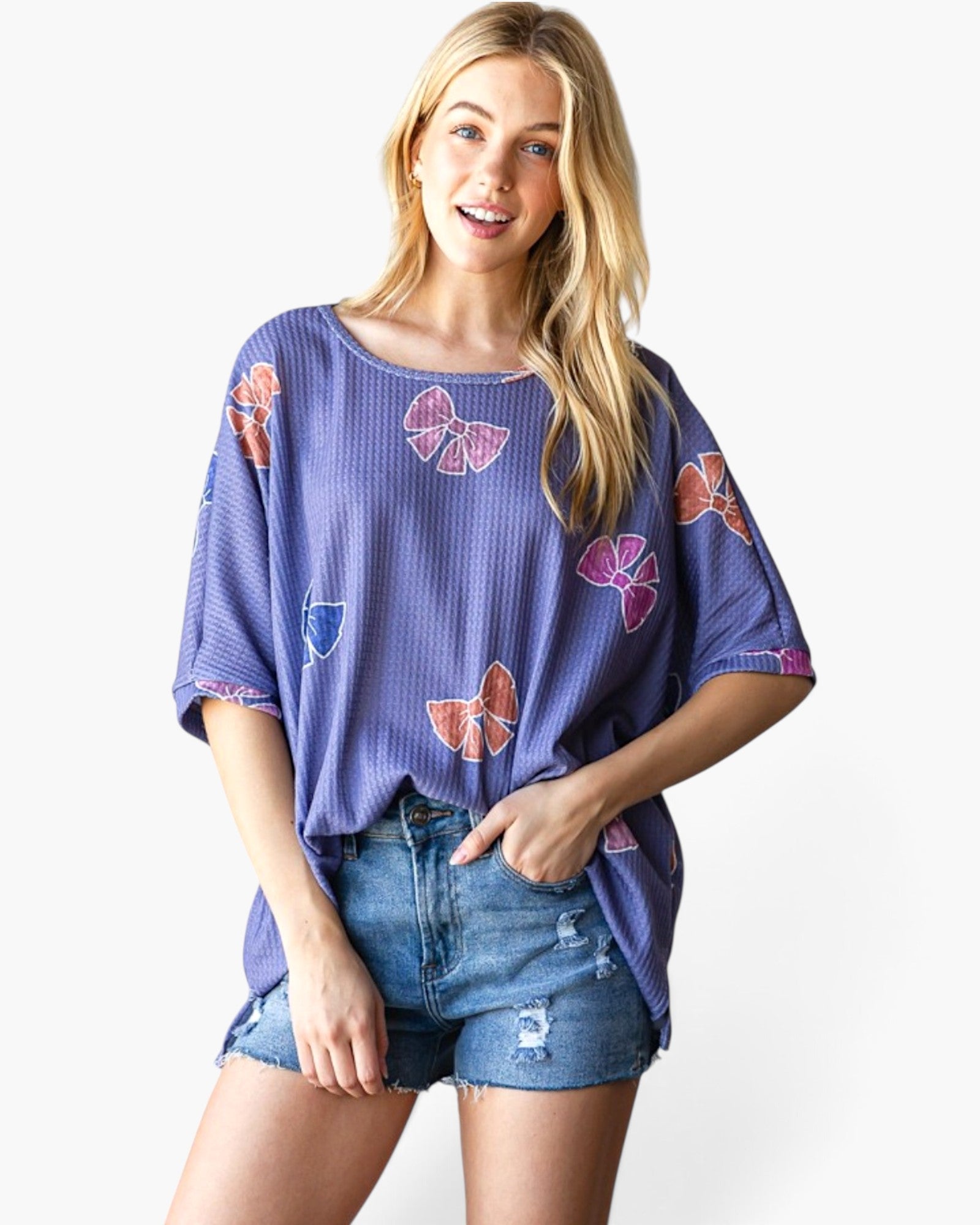 A woman modeling a purple waffle-knit short-sleeve top with colorful butterfly and bow prints, paired with distressed denim shorts, against a plain white background.