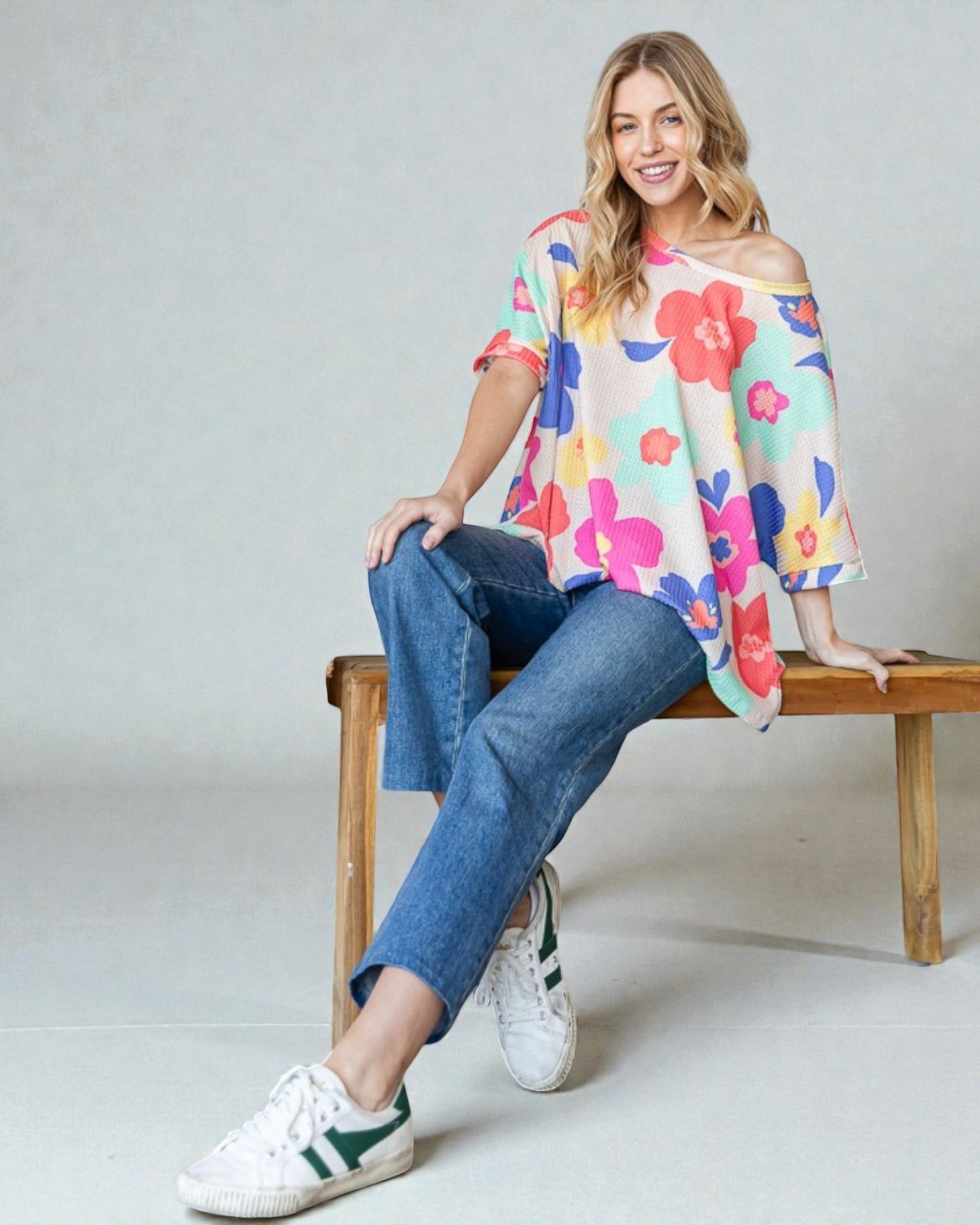 Woman sitting on a wooden bench wearing a colorful floral top and blue jeans.
