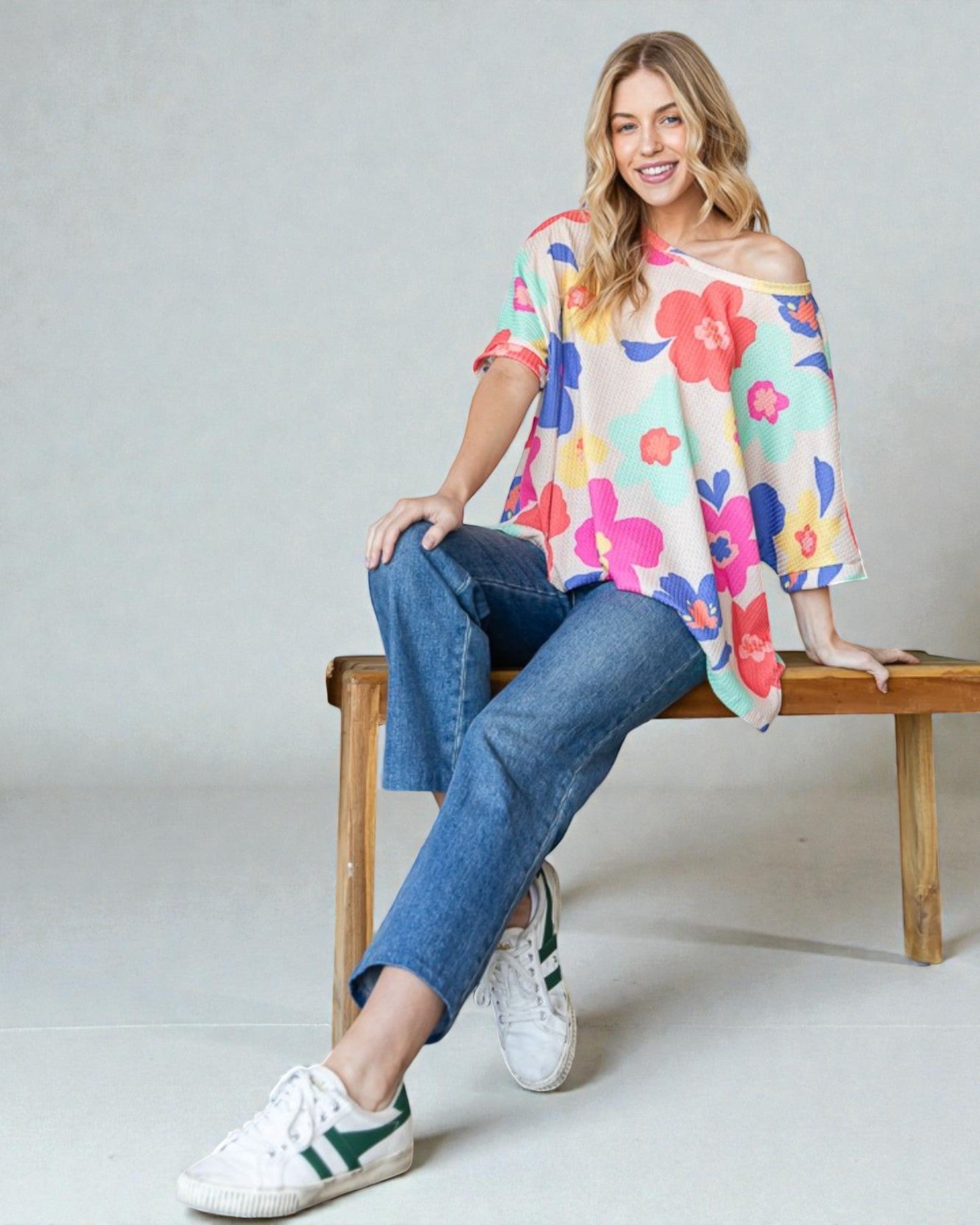 Woman sitting on a wooden bench wearing a colorful floral top and blue jeans.