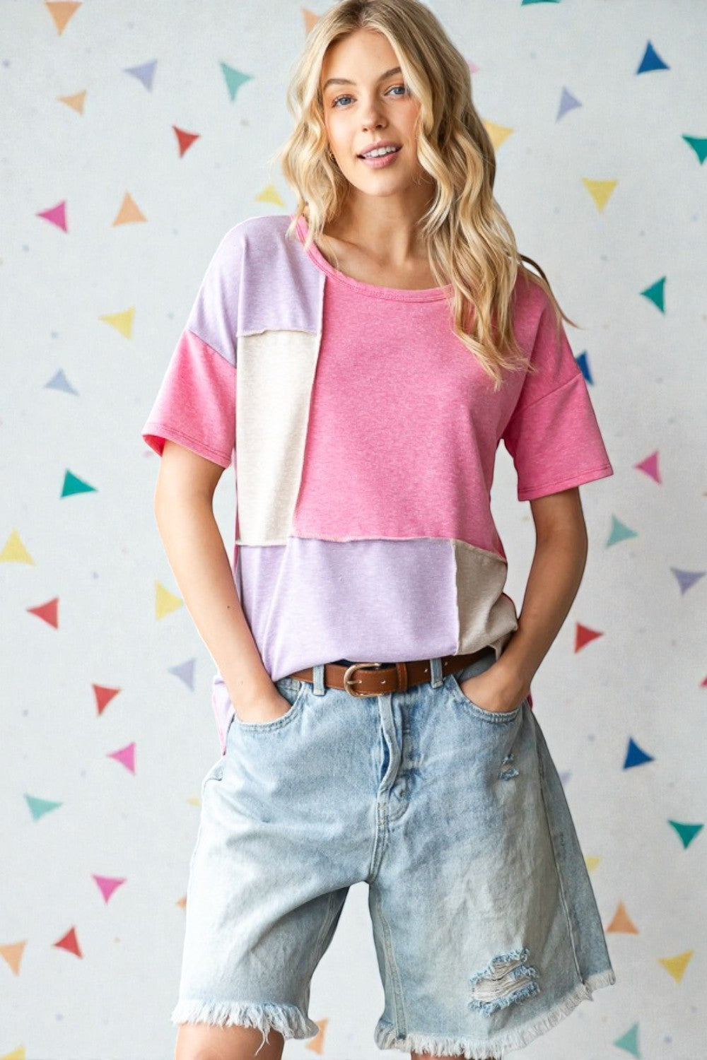 Woman wearing a colorful layered top and denim shorts against a multicolored triangle-patterned wall.