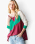 A woman wearing a color-blocked long-sleeve top with cream, green, and burgundy panels, paired with light-wash blue jeans, against a plain white background.