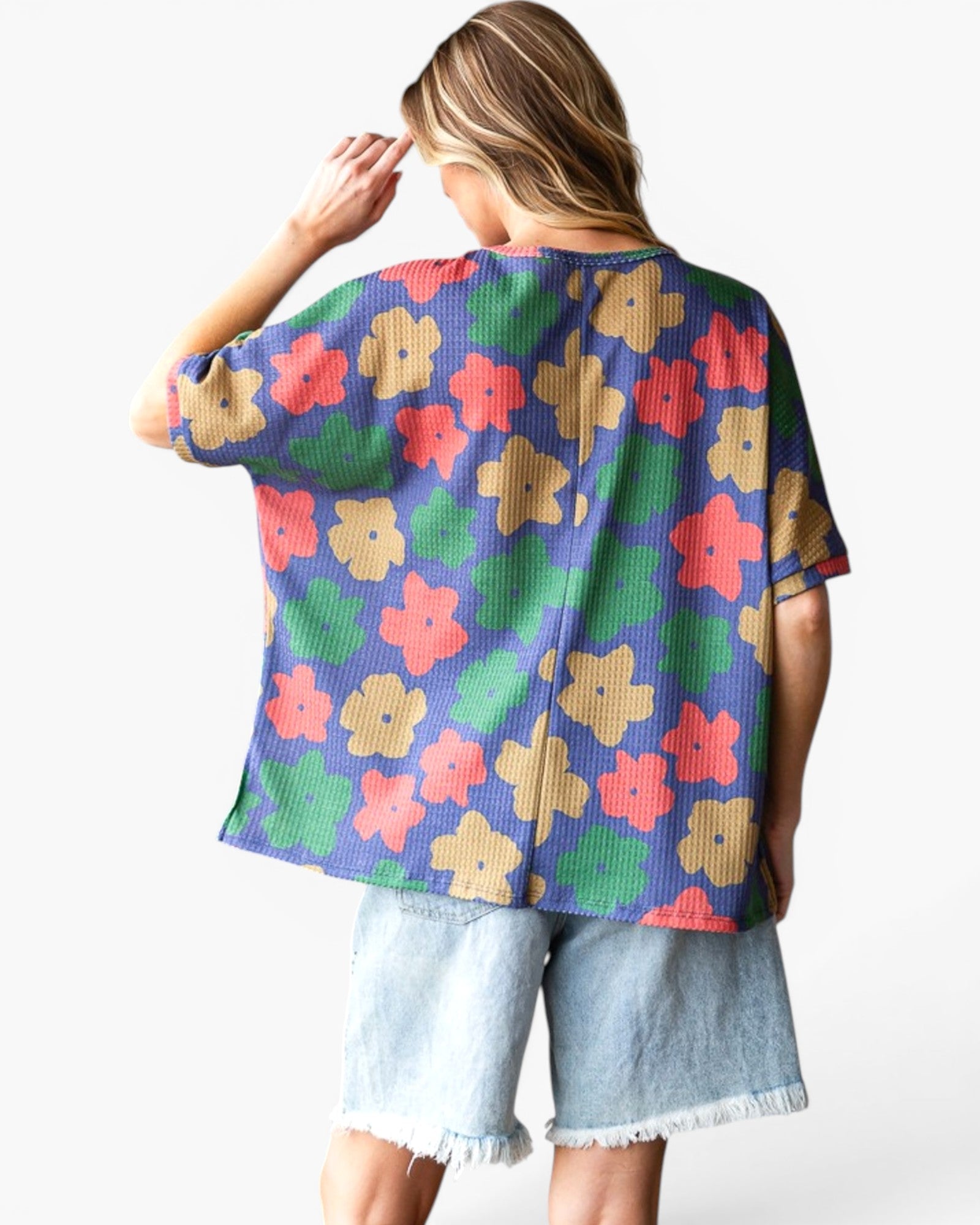 A woman shown from the back wearing a short-sleeve waffle-knit top with a colorful floral pattern in purple, green, red, and beige, paired with light-wash denim shorts against a plain white background.