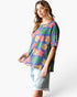 A woman modeling a short-sleeve knit top with a colorful floral pattern in purple, green, pink, and beige, paired with light-wash denim shorts against a plain white background.