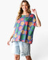 A woman modeling a short-sleeve waffle-knit top with a colorful retro floral pattern in purple, green, orange, and beige, paired with light-wash denim shorts against a white studio background.