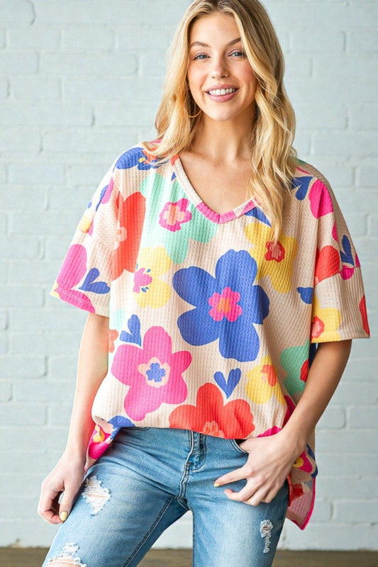 Woman wearing a colorful floral top and jeans against a white brick wall.