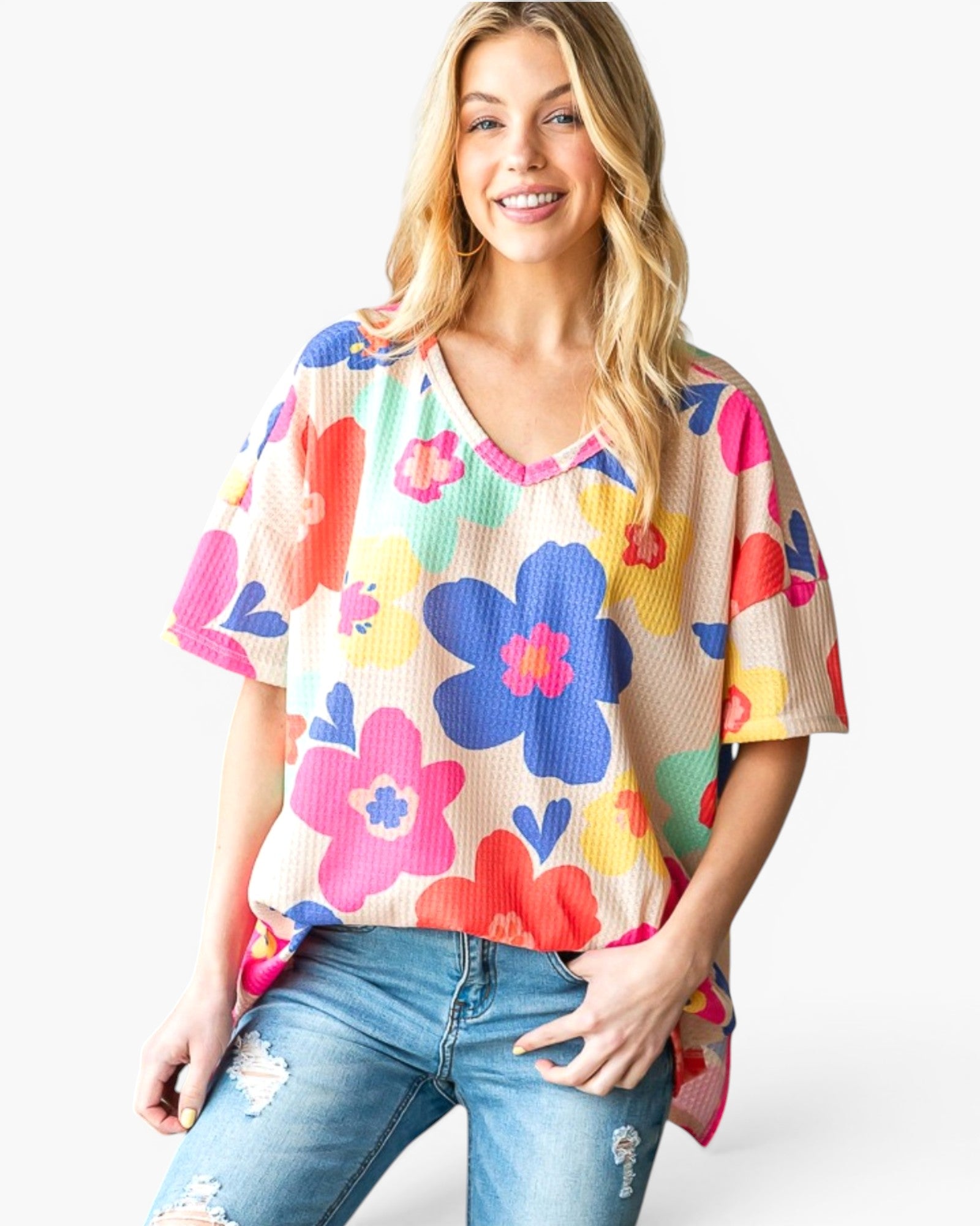 A woman wearing a colorful floral waffle-knit short-sleeve top and distressed blue jeans, standing against a light gray studio background.