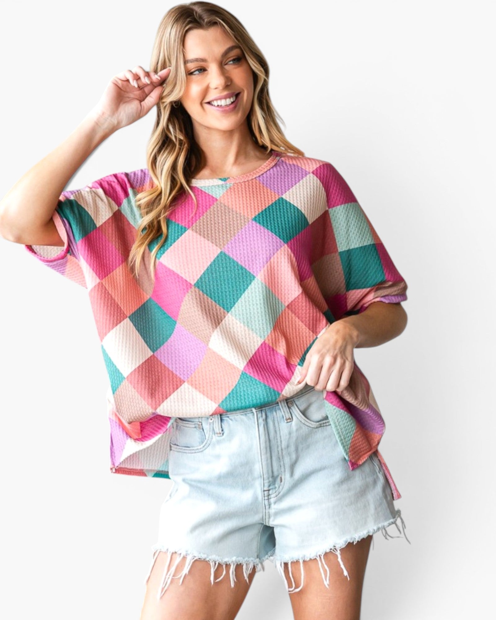 A woman modeling a short-sleeve, loose-fitting top with a colorful diamond argyle pattern in pink, teal, peach, and cream, paired with light-wash denim shorts against a plain white background.
