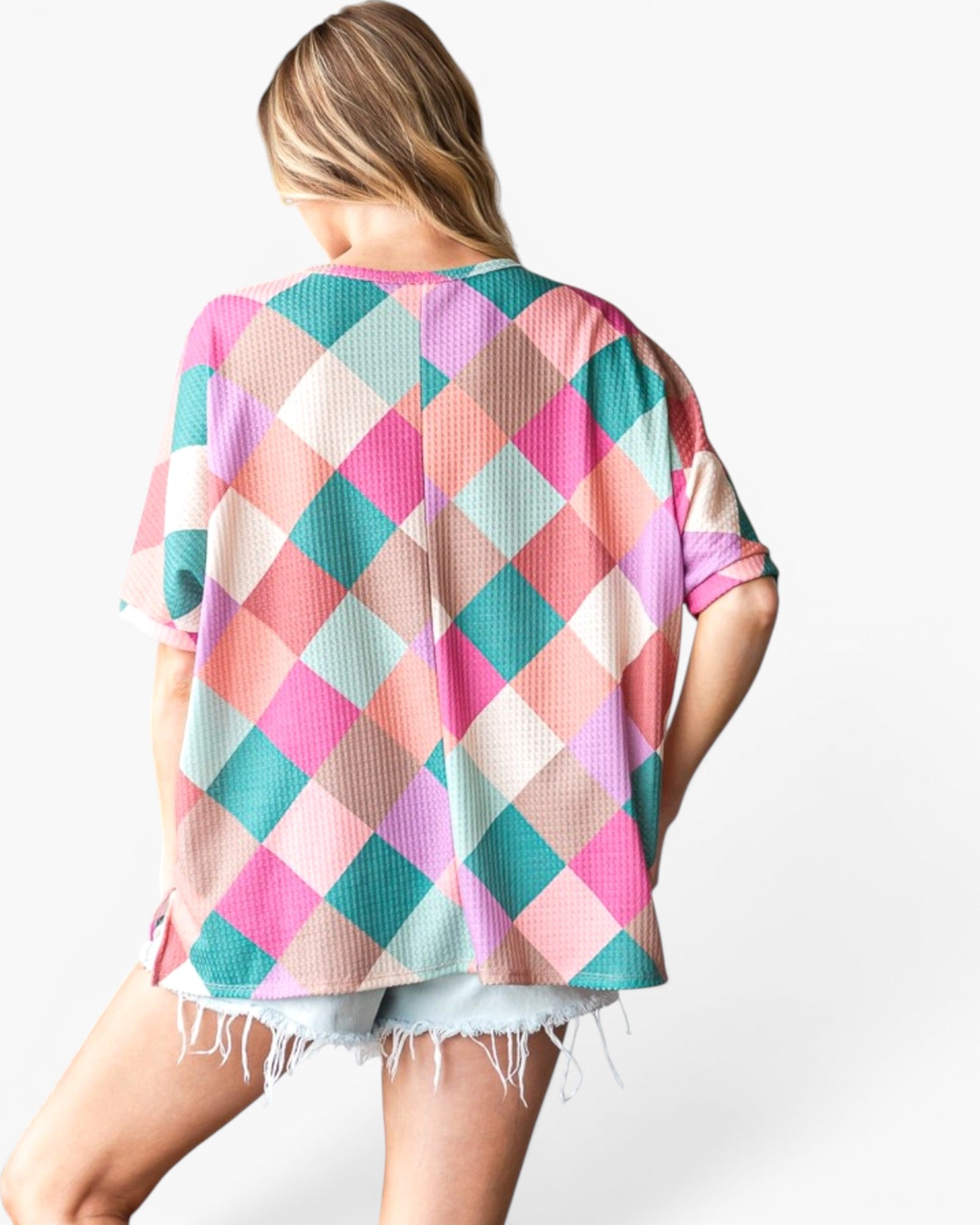 A woman shown from the back wearing a loose short-sleeve top with a colorful harlequin diamond pattern in pink, teal, peach, and cream, paired with light denim shorts against a plain white background.