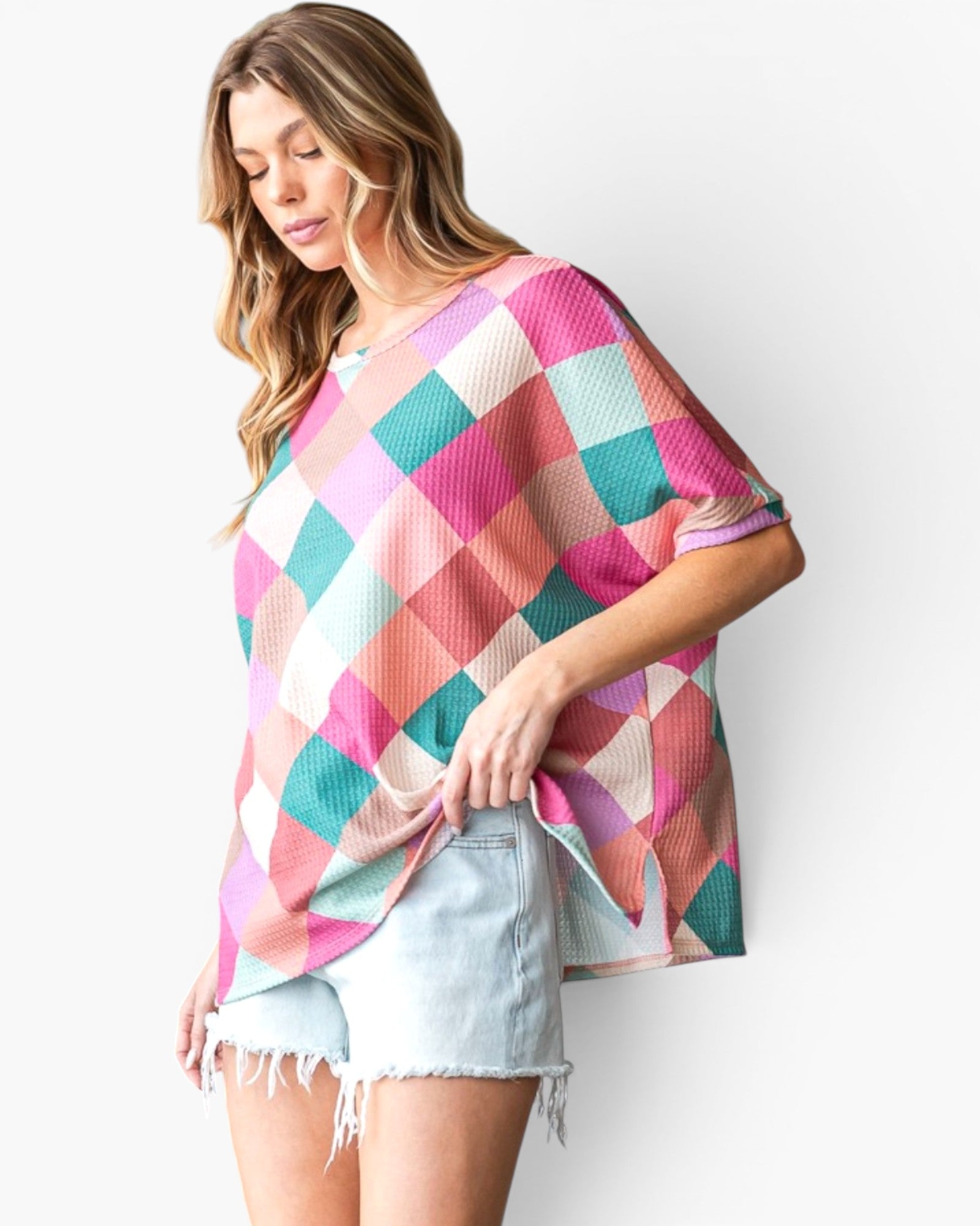 A woman wearing a loose, short-sleeve top with a colorful diamond pattern in pink, teal, peach, and cream, paired with light denim shorts, against a plain white background.