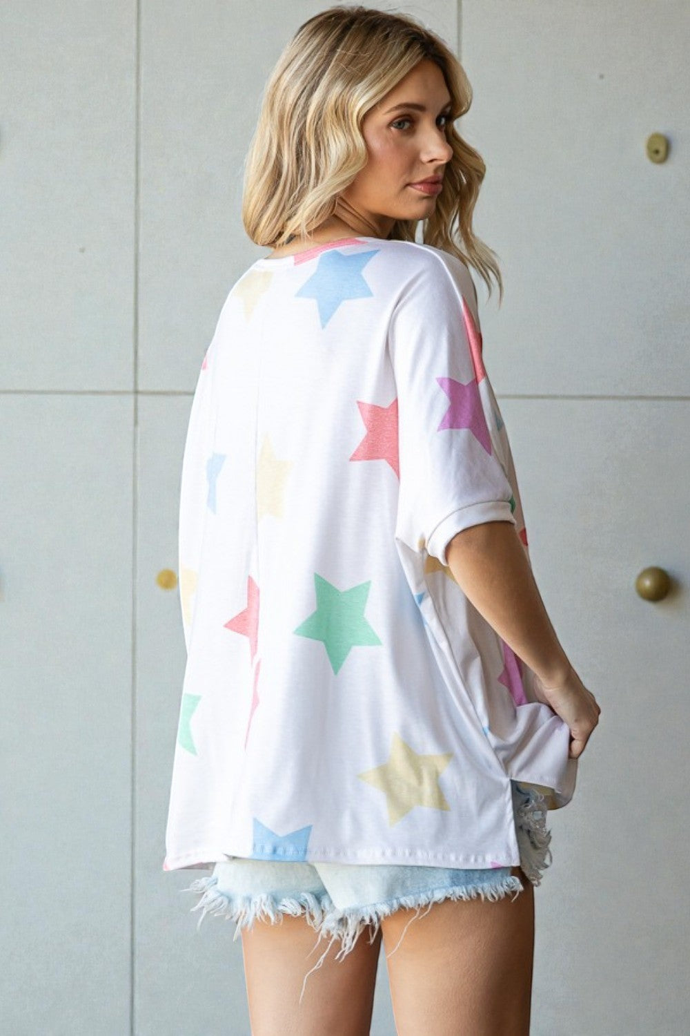 Woman wearing a white oversized t-shirt with colorful star patterns and denim shorts against a light-colored wall.
