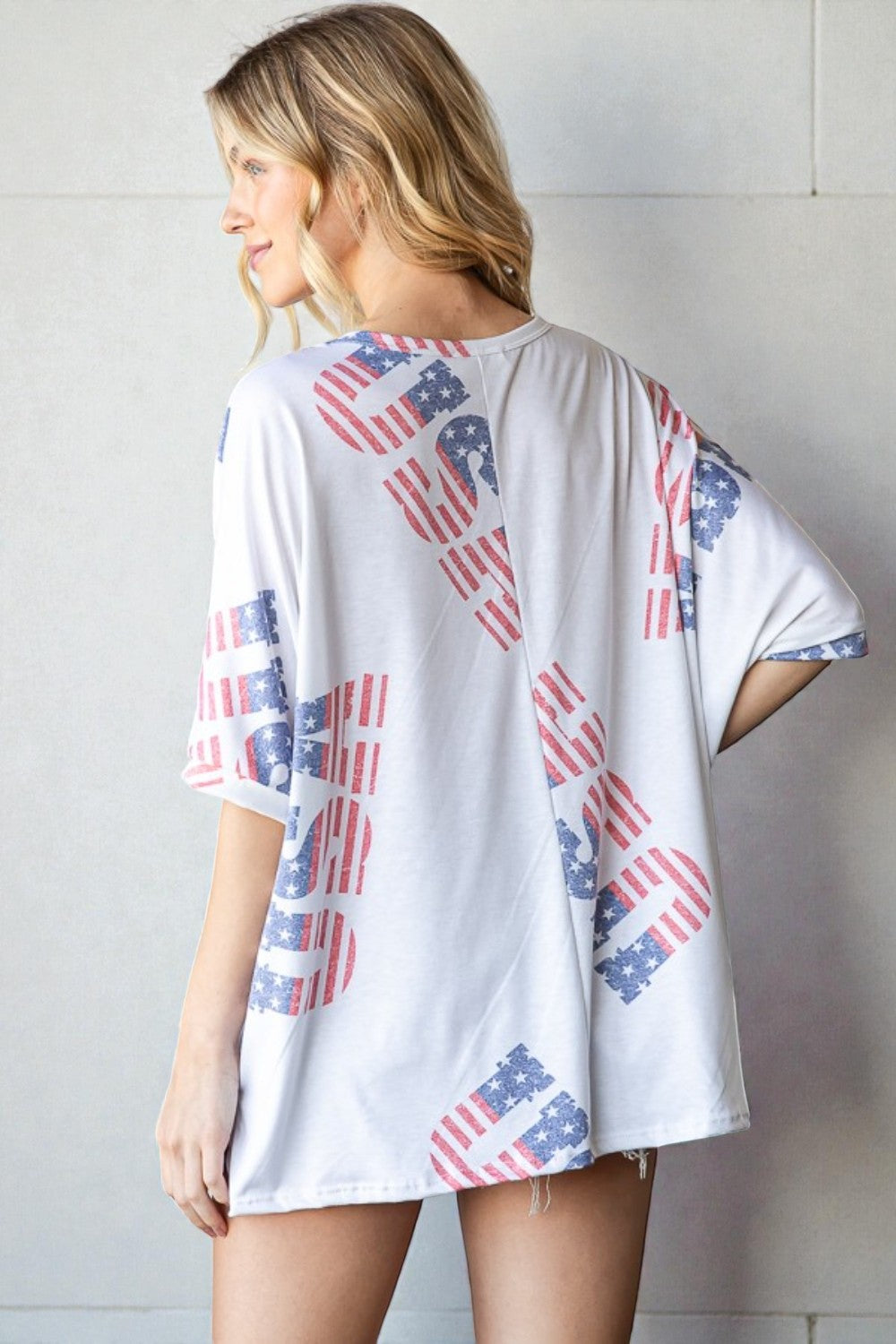 Woman wearing a white t-shirt with American flag patterns on the back and sleeves against a light gray wall.