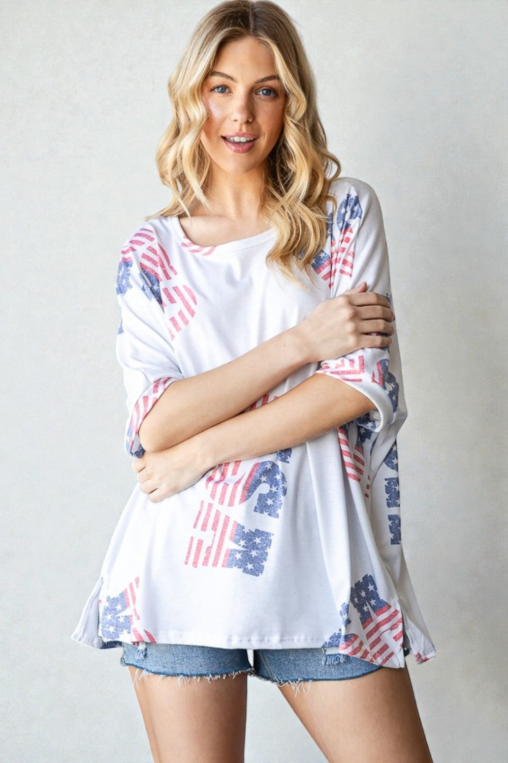 Woman wearing a white top with American flag pattern and denim shorts on a plain background