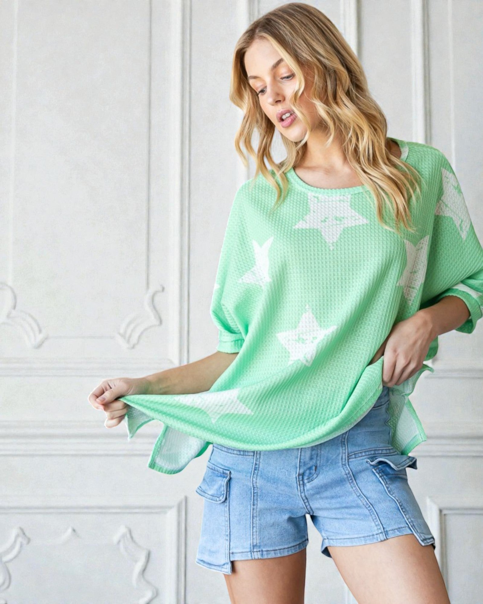 Woman wearing a light green sweater with star patterns and denim shorts against a white wall.