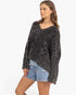 A woman modeling a dark charcoal gray cable-knit sweater with a V-neck and fringed hem, paired with light-wash denim shorts, against a plain white studio background.