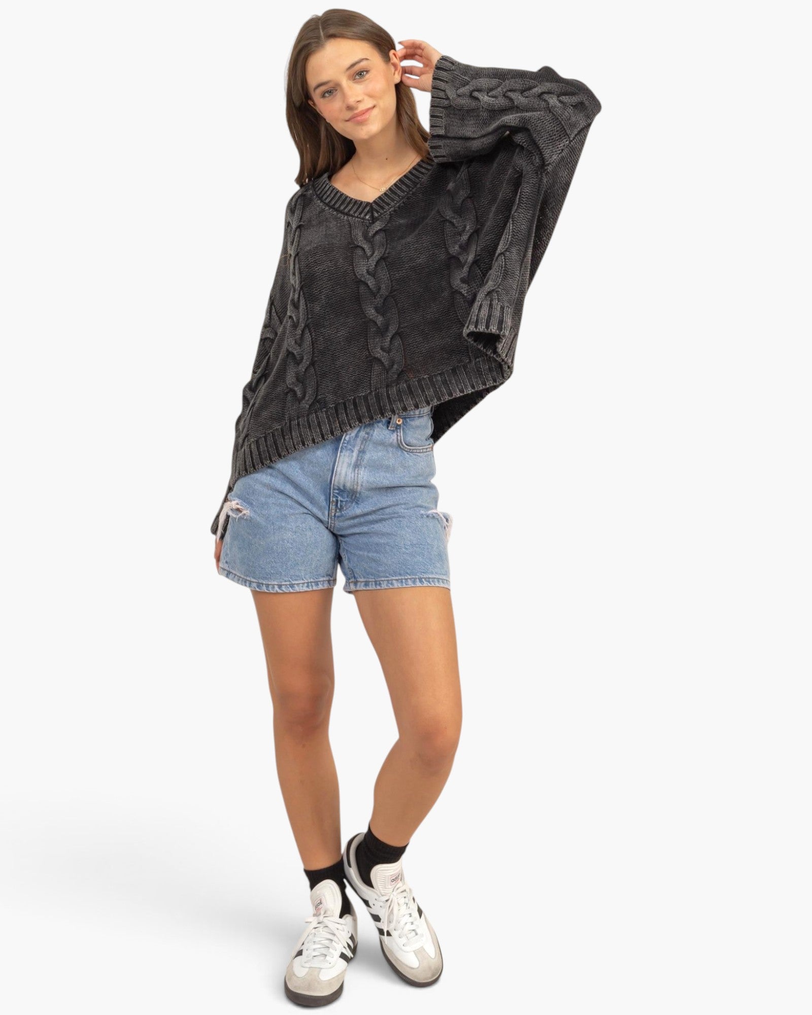 A woman modeling a dark gray washed cable-knit sweater, paired with light blue denim shorts, black socks, and white sneakers, against a plain white studio background.