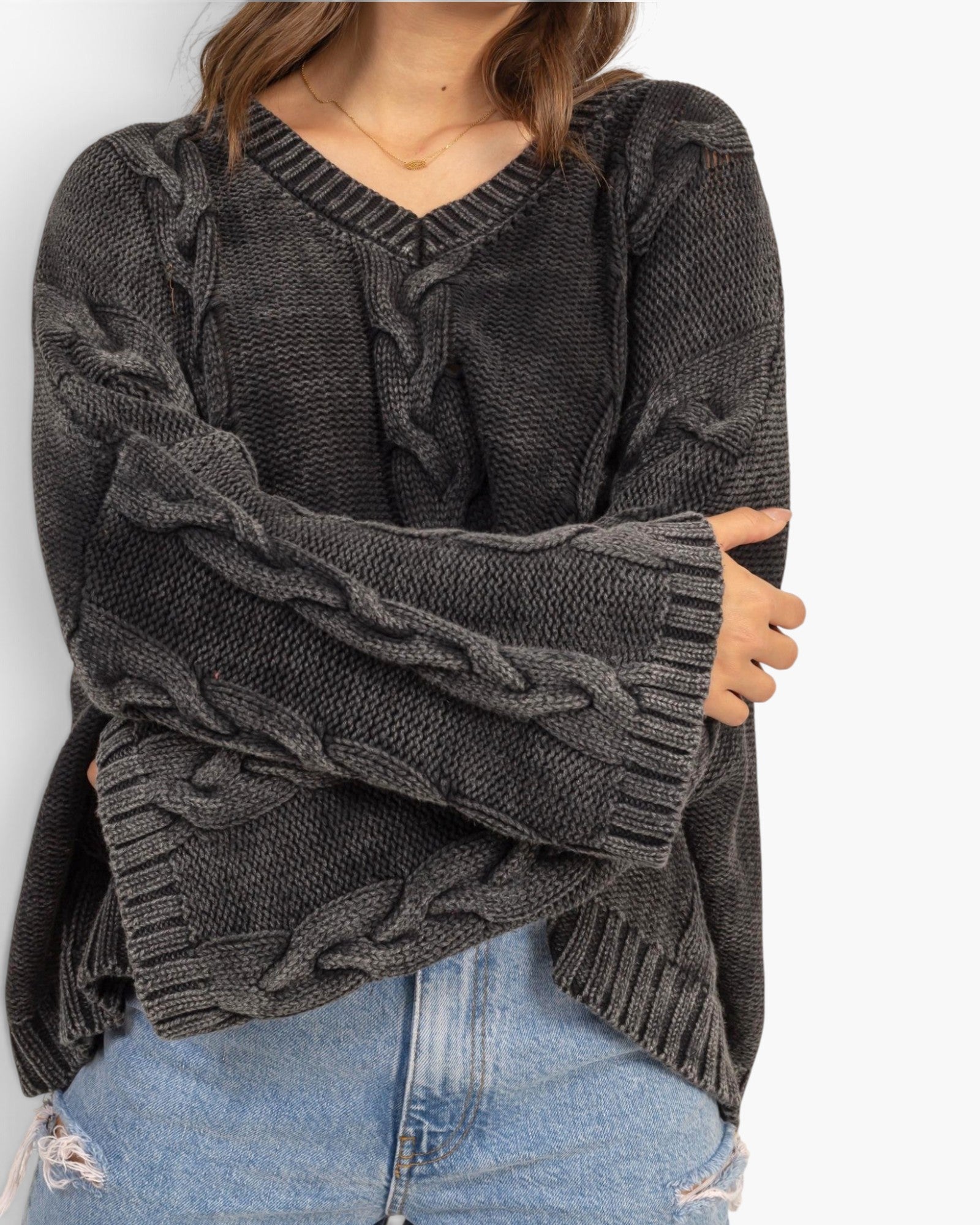A person wearing a dark charcoal gray cable-knit sweater with a V-neck and ribbed cuffs, paired with light blue jeans against a plain white background.