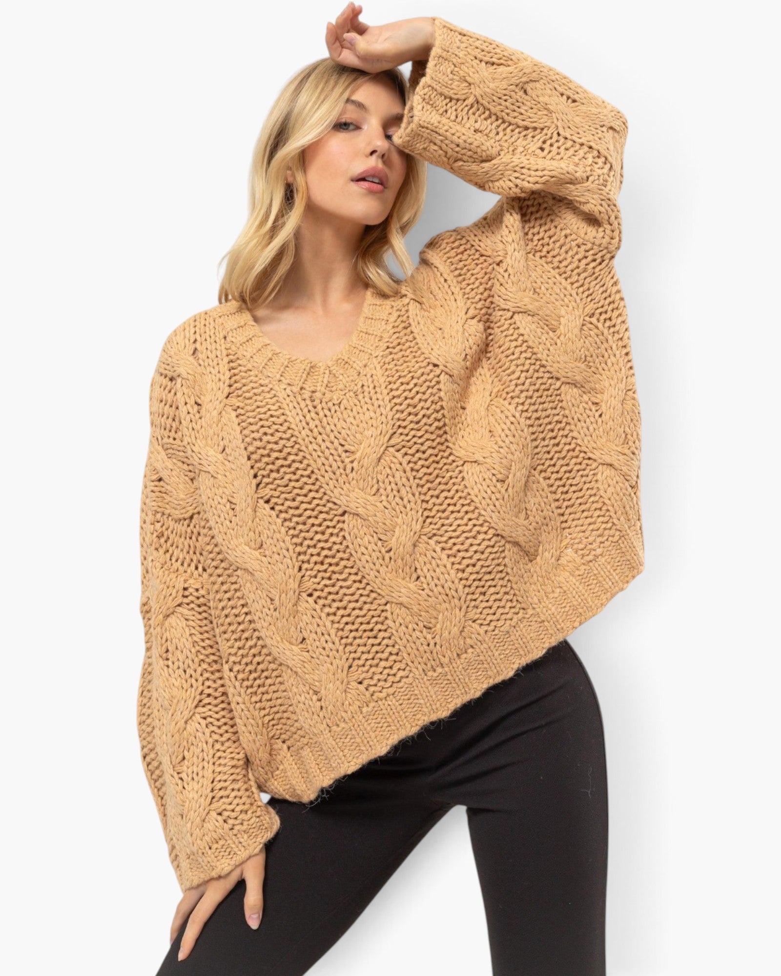 A woman wearing a chunky cable-knit cropped sweater in a warm camel tan color, paired with black pants, against a clean white background.