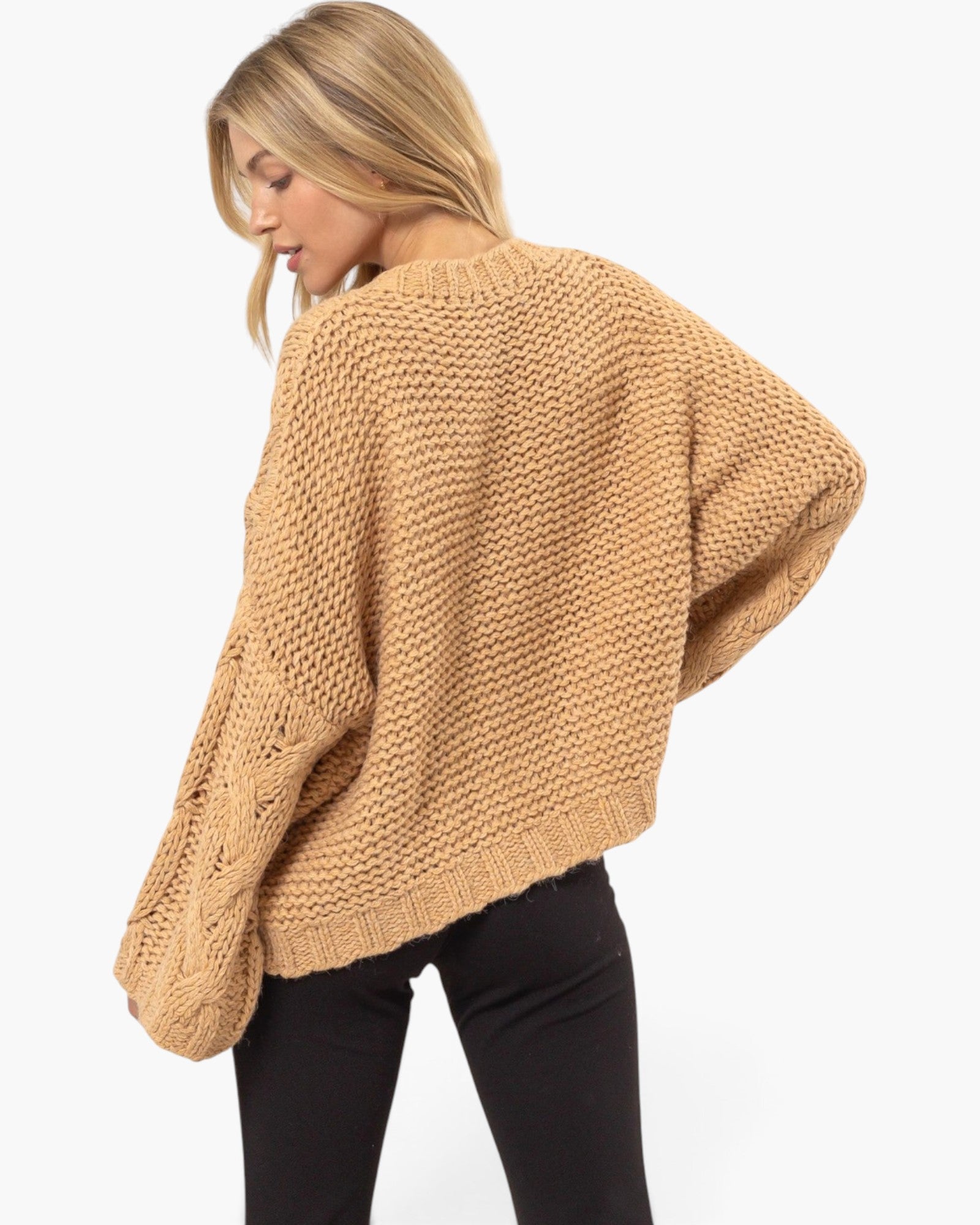 A woman wearing a chunky knit cropped sweater in camel tan, shown from a three-quarters back view against a plain white background.
