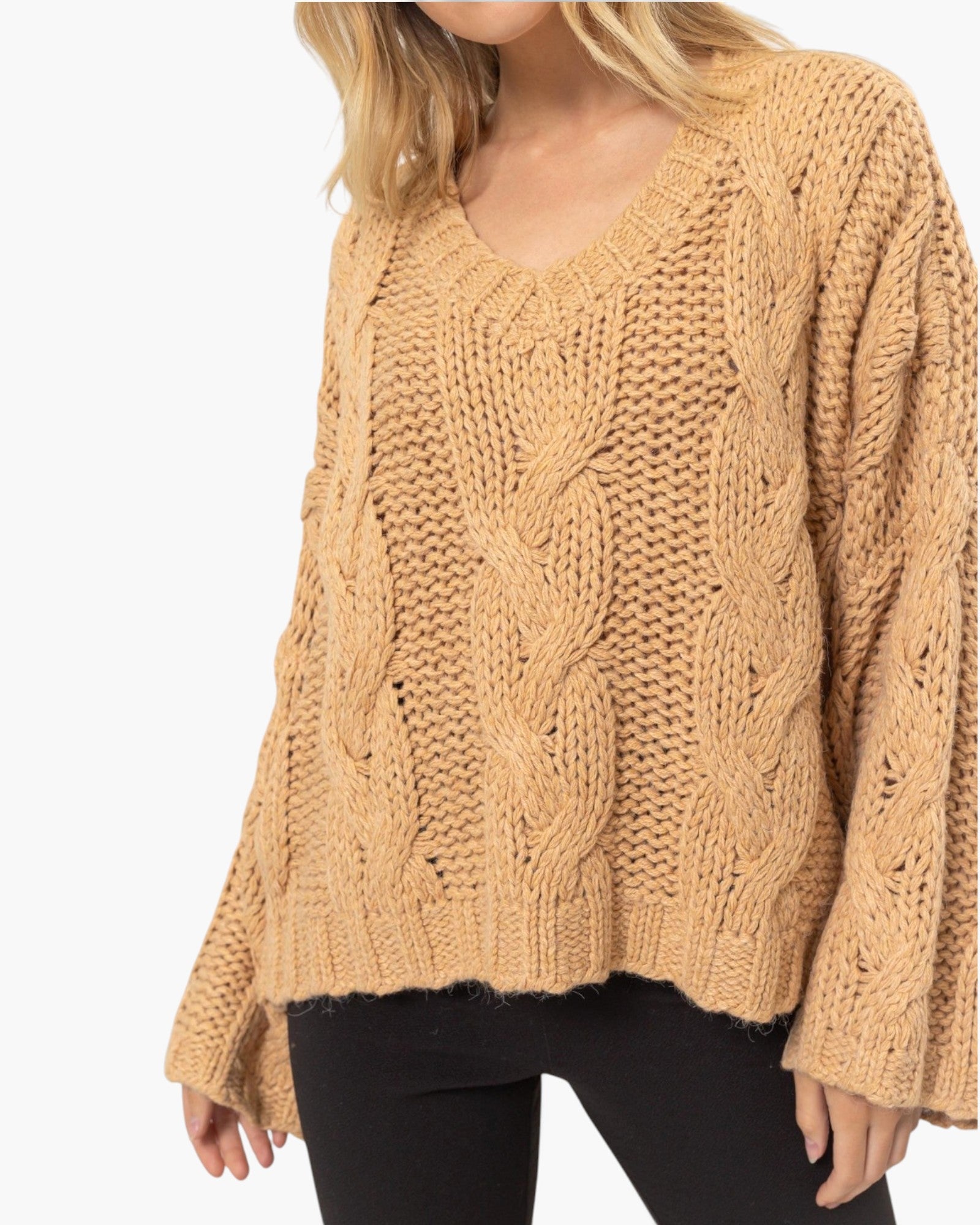 A woman wearing a chunky cable-knit sweater in a warm camel tan color, paired with black pants, against a plain white background.