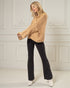 Woman wearing a beige knitted sweater and black pants against a white wall.