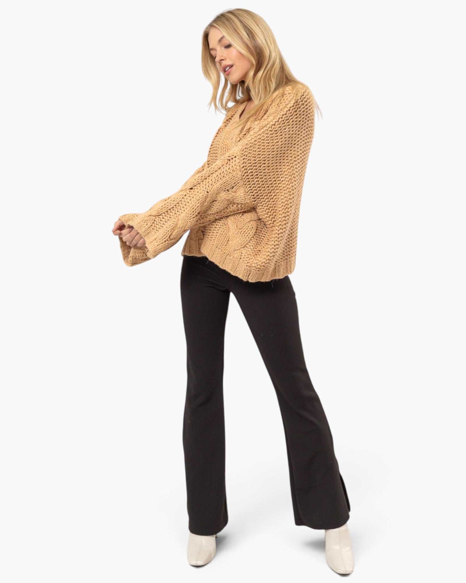 A woman modeling a chunky knit cropped sweater in camel beige with long bell sleeves, paired with black flared pants and off-white pointed shoes, against a plain white studio background.