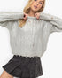 A woman wearing a chunky silver cable-knit sweater with distressed details and a dark gray pleated mini skirt, posing against a plain white background.