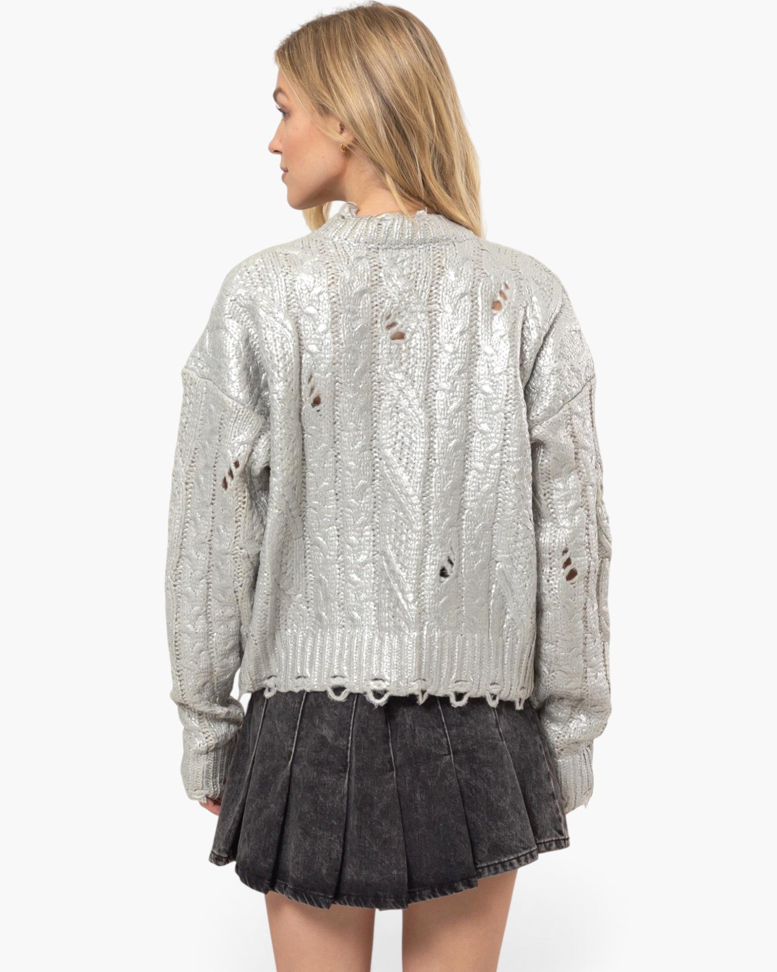 A woman shown from the back wearing a metallic silver cable-knit sweater with distressed holes and a dark gray pleated mini skirt, against a plain light gray studio background.
