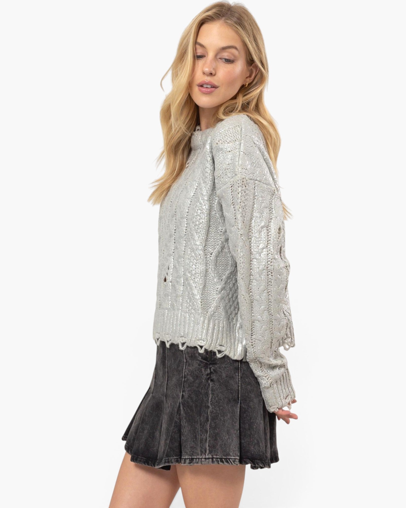 A woman modeling a light gray cable-knit sweater with distressed, frayed edges and a dark gray pleated mini skirt against a plain white studio background.