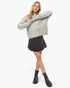 A woman modeling a chunky light gray cable-knit cropped sweater with frayed edges, paired with a dark gray pleated mini skirt and black ankle boots, against a plain white studio background.