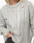 A close-up of a person wearing a chunky knit sweater with distressed holes. The sweater has a textured cable and floral knit pattern in a light silver-gray color.