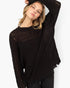 A woman modeling a black open-knit, oversized sweater with a crew neck and raw, frayed edges, paired with black pants against a plain white background.