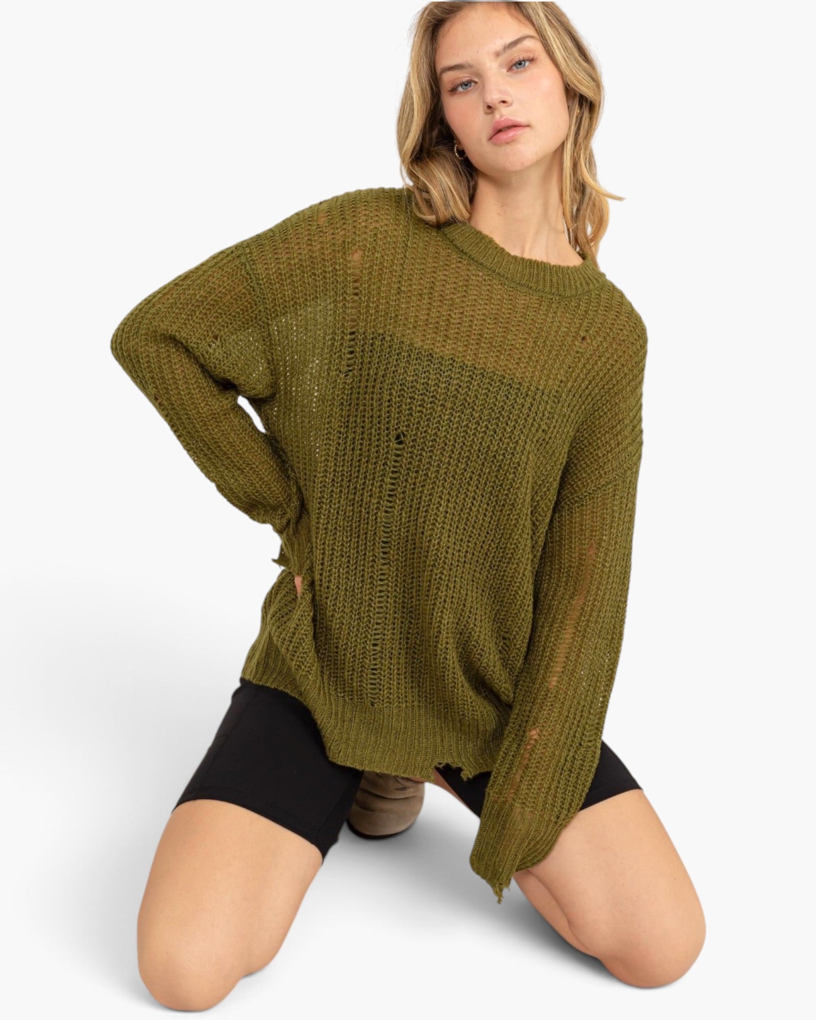 A woman kneeling on a light gray studio background wearing an oversized olive green distressed knit sweater and black shorts.