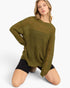A woman kneeling on a light gray studio background wearing an oversized olive green distressed knit sweater and black shorts.