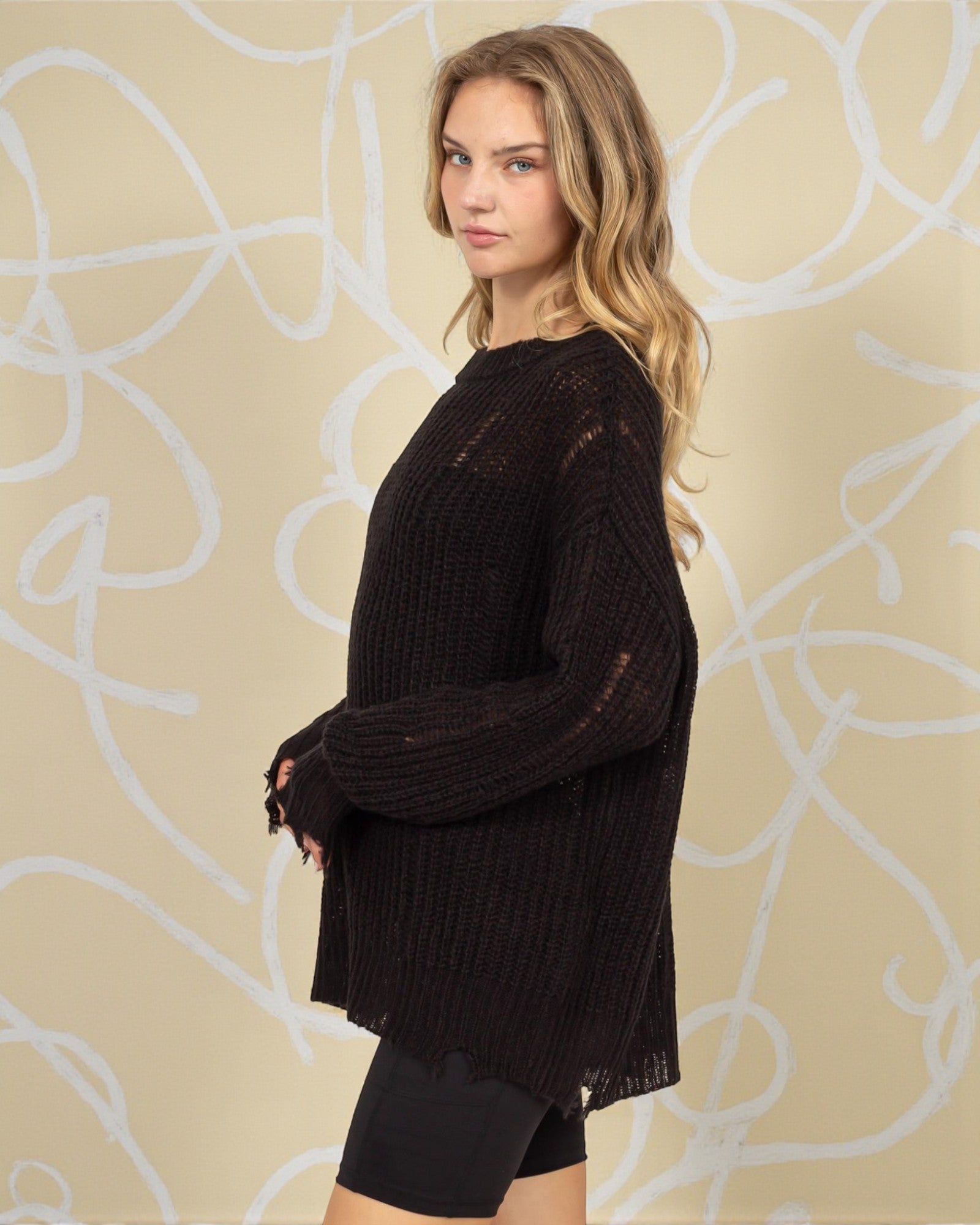 Woman wearing a black sweater and shorts against a beige wall with white abstract patterns