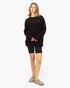 A woman modeling an oversized dark brown ribbed knit sweater with a sheer lace-like yoke, paired with black biker shorts and beige suede loafers, against a plain white studio background.