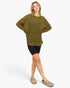 A woman modeling an oversized olive green ribbed knit sweater with distressed details, paired with black bike shorts and beige slip-on shoes, against a plain white studio background.