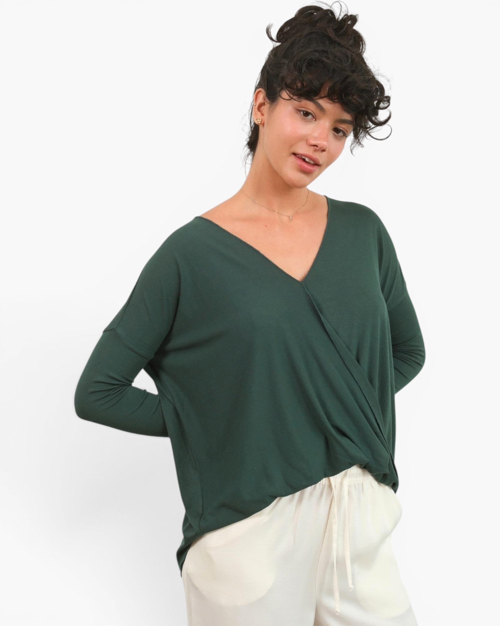 A woman wearing a dark green long-sleeve wrap-style top and white pants, photographed against a plain light gray studio background.