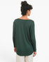 A woman viewed from the back wearing a dark green long-sleeve top with a wide boat neckline and cream pants, set against a plain white studio background.