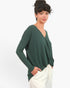 A woman modeling a dark green long-sleeve wrap-style top with a V-neck, paired with white pants, against a plain white studio background.