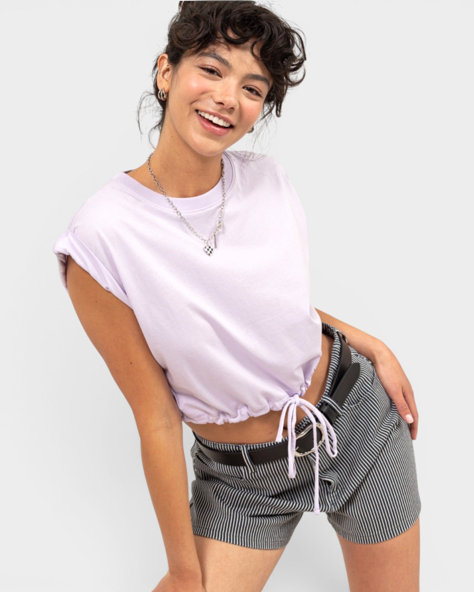 A woman modeling a light lavender short-sleeve crop top and black-and-white pinstriped shorts against a light gray studio background.