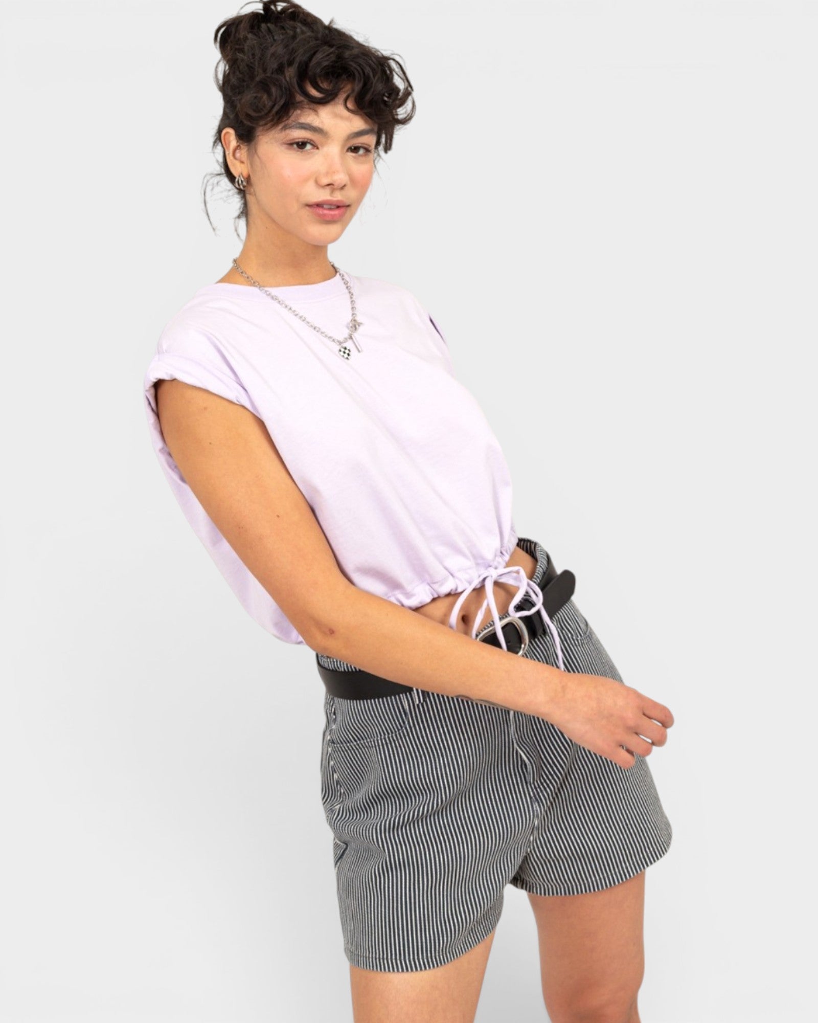A woman modeling a light lavender short-sleeve crop top and gray-and-white pinstriped shorts against a light gray studio background.
